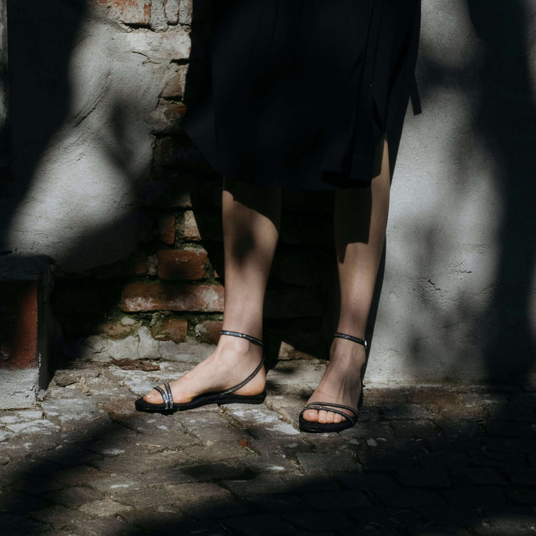 Model wearing Faye black crystal vegan flat sandals with thin crystal-embellished straps in an outdoor setting.